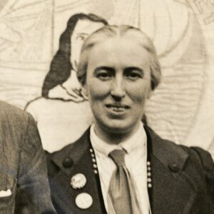 The portrait of Clemence Housman is a detail taken from a photograph published in The Evening Standard at the time of her arrest for tax evasion as a form of suffragist protest. Housman is looking directly at the camera and smiling. Her hair is pulled back and she is wearing a dark jacket, white shirt, and tie. She has suffrage badges on her lapel and is standing in front of a suffrage banner.