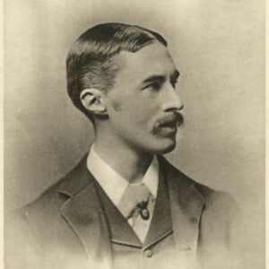 This is a sepia photograph of A. E. Housman when he was 35 years old. Housman’s face is in profile facing right and his body is turned towards centre. He is wearing a dark coloured dress jacket, a similarly coloured buttoned up vest, a collared shirt, and puff tie with a pin. Housman has short dark hair parted in the centre of his head and slicked down. He has medium length sideburns and a moustache with the corners extending just below the corners of his lips. Housman’s mouth is slightly opened.