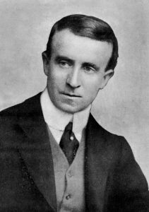 John Buchan is shown from the middle of his chest up, his face turned towards the right, eyes also turned towards the right. He is wearing a suit, vest, tie, and high-collared shirt. The background is light-coloured.