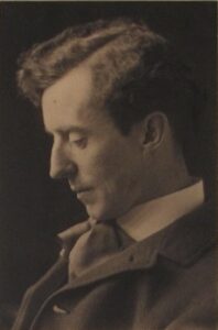 The sepia coloured photograph shows a head-and-shoulders profile view of a young Charles Dalmon, looking down. He is clean-shaven and has thick wavy hair parted on the side. He is wearing a dark jacket with high white colour and loose tie.