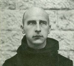 The tonal black-and-white photograph is a head-and-torso shot of a middle-aged Benedictine monk in a black cowl and shaved head, standing in front of a brick wall.