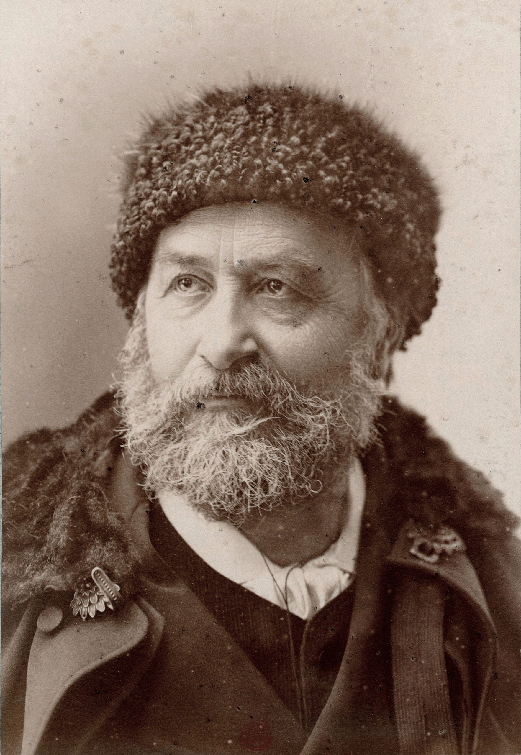 The sepia photograph of Élie Reclus is a head-and-shoulders shot in three-quarter view of an older man with a grizzled beard and moustache, wearing a Persian wool hat and a coat with a fur collar.