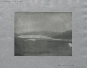 This half-tone reproduction is mounted on special grey art paper, unframed, centered on the page in landscape orientation. The image depicts a landscape with a lake, surrounded by trees and hills. Printed below the image is: “THE LAKE AT COOLE. / From a pastel by W. B. YEATS.”