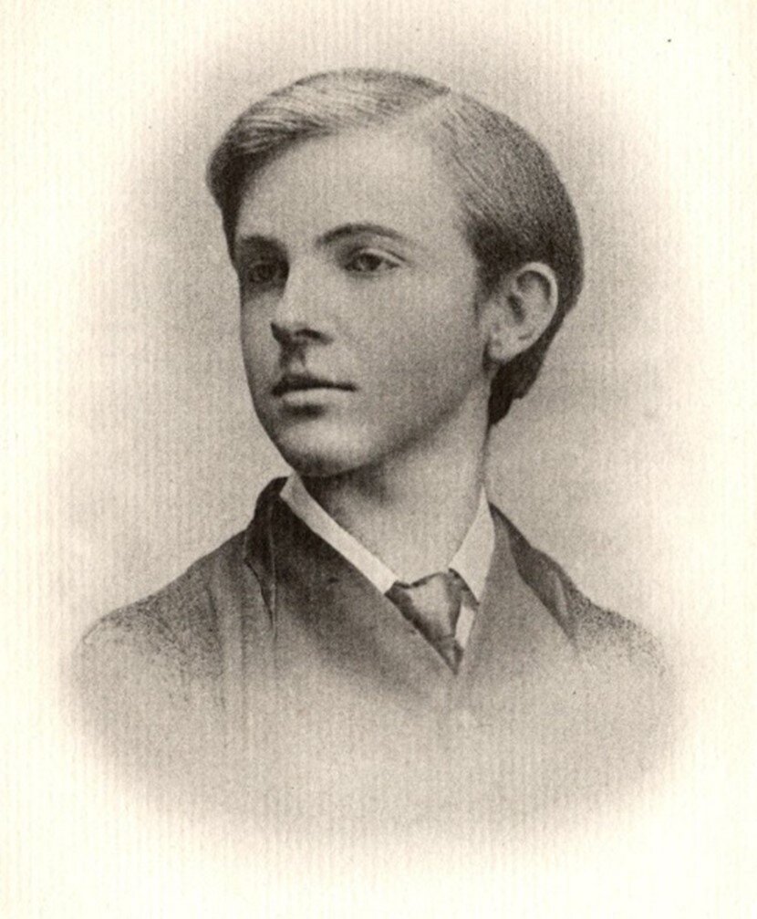 This is a sepia photograph portrait of Lionel Johnson at the age of 18. Johnson’s body can be seen from the bottom of his shoulders to his head. His body is centred while his face and gaze are angled to the left side of the image so his face shows in ¾ profile. Johnson is wearing a dark jacket, a dark vest, a light collared shirt, and dark neck tie. His hair is short and neatly combed; the part in his hair is off centred.