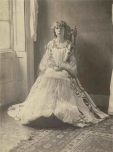 The tonal photograph shows a young woman with bangs and jaw-length hair, seated in a carved chair by a window, and facing the camera directly. Her hands are folded on her lap and her feet are resting on a footstool. She is wearing a long white dress and a long cape decorated with lace, applique, and embroidery.
