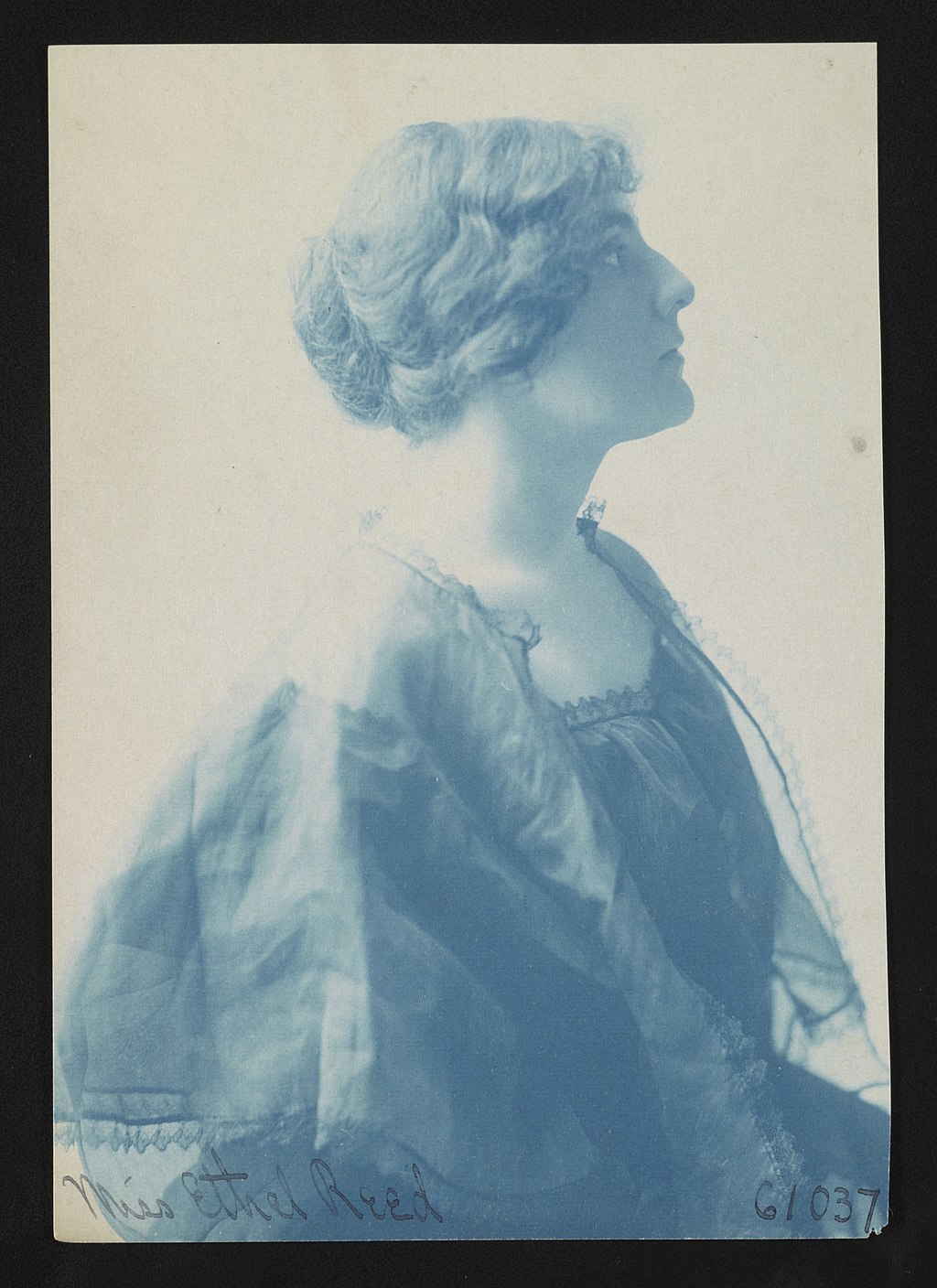 The gray-toned photograph shows Ethel Reed’s head and torso in profile, facing right. Her abundant hair is twisted back into a chignon; her face in profile is unsmiling. She is wearing a dark dress with a square neckline lined with lace. A semi-transparent cape with a lace edging covers her shoulders. A handwritten inscription at the bottom of the photograph reads: Miss Ethel Reed 61037.