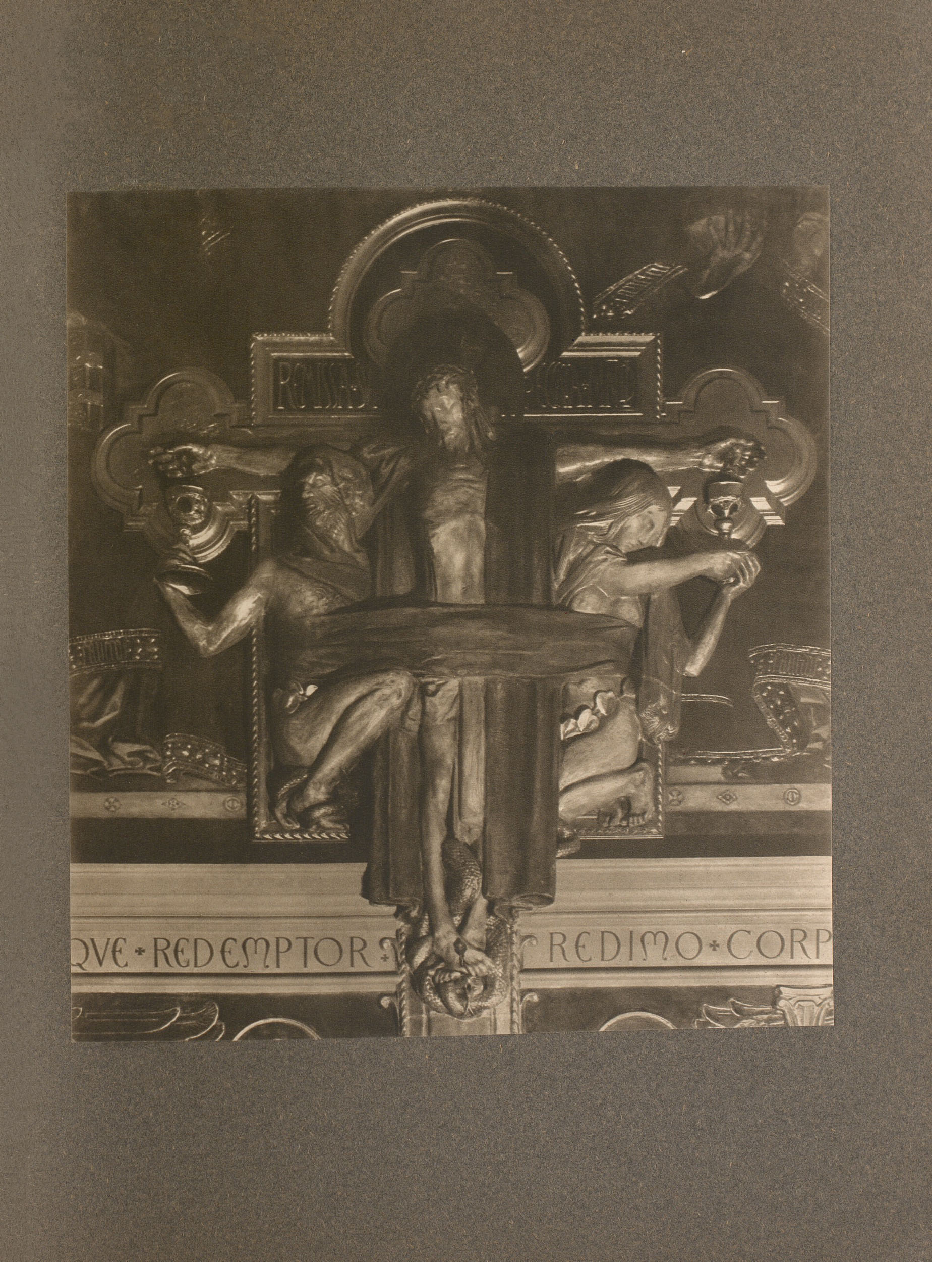 The tonal image is in portrait orientation and is located in the center of the page, tipped in on dark art paper. The photogravure of the relief sculpture presents a darkened image of the crucifixion, symbolically representing redemption by including the figures of Adam and Eve at the base of the cross. Christ is on the cross, with his head tilted downward, toward Adam and Eve on either side of him below.. There is a dark garment around the back of Christ’s’ neck that rests on either side of his body. Another dark garment is wrapped around Christ’s waist, binding him to Adam and Eve. On the left, Adam is crouched by the foot of the cross, with a serpent around his feet; he holds a chalice up to Christ to catch his blood. Eve is crouched at the foot of the cross on the right of Christ, wearing a belt made out of leaves and holding a chalice up to catch Christ’s blood. Below the image on the wall behind it is the Latin inscription: “AVE, REDEMPTOR, REDIMO, CORP.”
