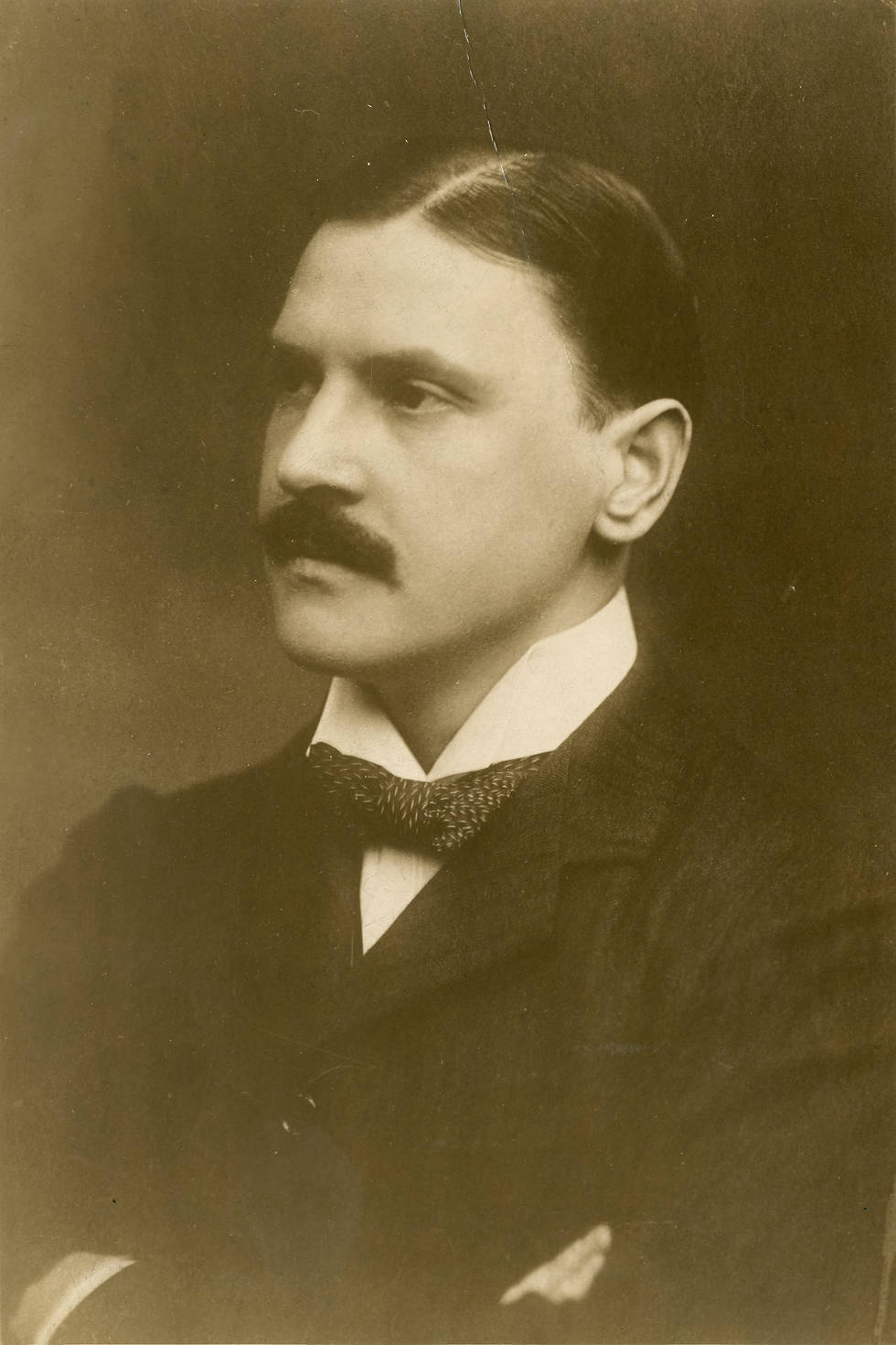 The sepia-toned photograph is a head-and-shoulders portrait of Somerset Maugham at age 37. He is posed in three-quarter profile looking to the left, with his arms folded across his chest. His dark hair is parted near the middle and brushed smoothly back. He has a dark bushy moustache. He is wearing a stiff white collar, bow tie, and dark jacket.