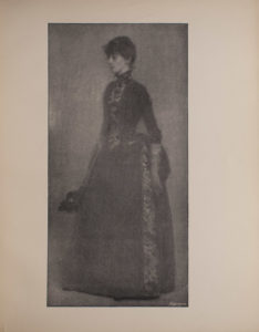 The image is of a woman standing and turned to the left She is wearing a dark coloured dress with a panel of pattern running vertically along the front of the bodice and the side of the skirt of the dress The image is vertically displayed