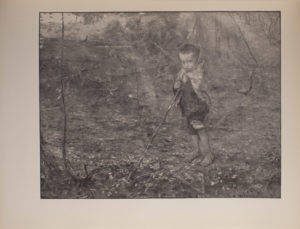 The image is of a young boy in a wooded area The boy stands barefoot holding a long stick that extends diagonally to the left He is wearing pants which are rolled up to his knees and appears to have a scarf or blanket wrapped around his shoulders and the back of his head The boy faces the viewer The image is horizontally displayed