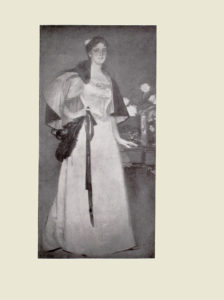 Image is of a woman shown full frontally Her teeth are visible through her slightly parted teeth She is wearing a long detailed gown that reaches the floor Only her left foot can be seen it sticks out slightly from underneath her dress Her hair is tied back The a line dress has a fitted bodice She is wearing a short dark cape part of the cape is draped around her chest. The womans right hand is holding on to a dark coloured ribbon and a dark feather her left hand is resting on a table The table divides the image horizontally The table has a vase of white flowers and a teacup and saucer The background is dark and undefined The image is vertically displayed