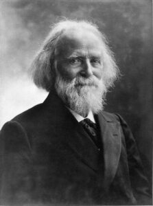 The black-and-white photographic portrait shows the head and shoulders of the elderly male subject, Élisée Reclus, in three-quarter profile. He has a receding hair line, long white hair, and a white beard and moustache. He is looking left toward the camera and smiling with closed lips. He is wearing a dark suit jacket with a white collared shirt and a dark tie.