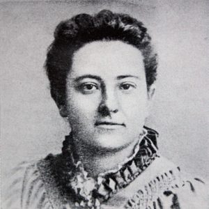 Black-and-white headshot of Olive Schreiner shows her from the shoulders up and facing the camera. Her eyes are gazing very slightly to her left. She is wearing a light-coloured dress or shirt with a dark-coloured, high, ruffled collar.